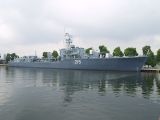 A large naval ship with the number 215 painted on its side is moored along a waterfront. The vessel is equipped with various antennas and radar equipment, and several trees are visible in the background, indicating a dock or port area. The water reflects the image of the ship, creating a calm and orderly scene under a cloudy sky.