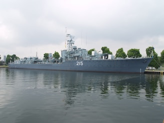 A large naval ship with the number 215 painted on its side is moored along a waterfront. The vessel is equipped with various antennas and radar equipment, and several trees are visible in the background, indicating a dock or port area. The water reflects the image of the ship, creating a calm and orderly scene under a cloudy sky.