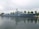 A large naval ship with the number 215 painted on its side is moored along a waterfront. The vessel is equipped with various antennas and radar equipment, and several trees are visible in the background, indicating a dock or port area. The water reflects the image of the ship, creating a calm and orderly scene under a cloudy sky.