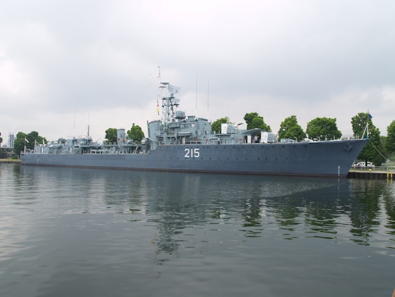 A large naval ship with the number 215 painted on its side is moored along a waterfront. The vessel is equipped with various antennas and radar equipment, and several trees are visible in the background, indicating a dock or port area. The water reflects the image of the ship, creating a calm and orderly scene under a cloudy sky.