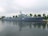 A large naval ship with the number 215 painted on its side is moored along a waterfront. The vessel is equipped with various antennas and radar equipment, and several trees are visible in the background, indicating a dock or port area. The water reflects the image of the ship, creating a calm and orderly scene under a cloudy sky.