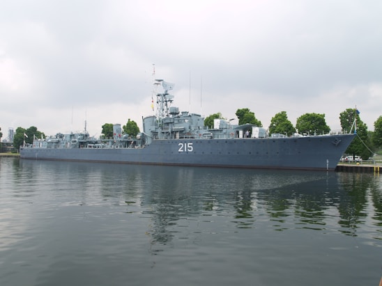 A large naval ship with the number 215 painted on its side is moored along a waterfront. The vessel is equipped with various antennas and radar equipment, and several trees are visible in the background, indicating a dock or port area. The water reflects the image of the ship, creating a calm and orderly scene under a cloudy sky.