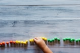 A child's hand places small colorful objects in a line on a wooden surface. The objects are arranged in sections by color, including red, yellow, and green.