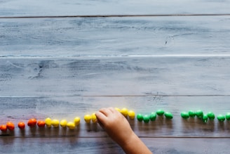 A child's hand places small colorful objects in a line on a wooden surface. The objects are arranged in sections by color, including red, yellow, and green.