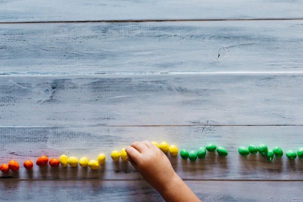 A child's hand places small colorful objects in a line on a wooden surface. The objects are arranged in sections by color, including red, yellow, and green.
