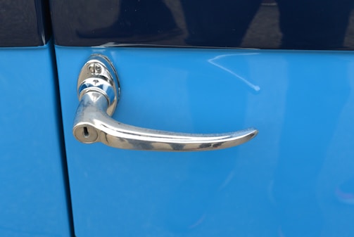 A shiny chrome car door handle attached to a blue vehicle. The handle has a sleek, rounded design with a keyhole below, mounted on a glossy surface.