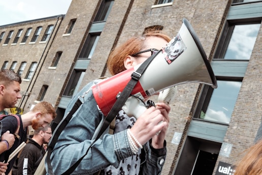 A group of people is gathered outside a brick building, one person holding a megaphone. This individual appears to be speaking or participating in a protest or public demonstration. The group seems focused and intent, suggesting an organized event.