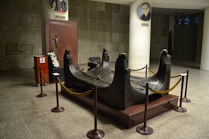A mausoleum with a bronze sculpture surrounded by rope barriers. The sculpture features four figures with solemn expressions, resembling mourning figures in a funeral setting. A red plaque with a crucifix is visible in the background, alongside floral arrangements. The area is clean and well-lit, with architectural elements such as tiled floors and white columns displaying portraits.