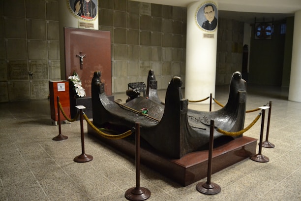 A mausoleum with a bronze sculpture surrounded by rope barriers. The sculpture features four figures with solemn expressions, resembling mourning figures in a funeral setting. A red plaque with a crucifix is visible in the background, alongside floral arrangements. The area is clean and well-lit, with architectural elements such as tiled floors and white columns displaying portraits.