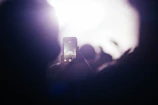 A person is capturing a photograph or video with a smartphone during a live music performance. The screen of the phone displays a stage with bright lights and performers. The background is filled with a hazy glow and silhouettes of the crowd.
