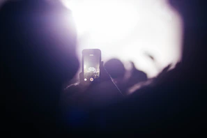 A person is capturing a photograph or video with a smartphone during a live music performance. The screen of the phone displays a stage with bright lights and performers. The background is filled with a hazy glow and silhouettes of the crowd.