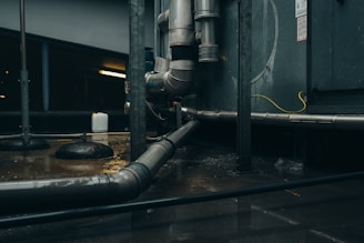 A dimly lit industrial setting with large metal pipes and ducts running across the scene. The area appears wet, with water on the floor and reflections visible. A small white container sits on the ground in the background, and some concrete pillars are present.