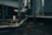 A dimly lit industrial setting with large metal pipes and ducts running across the scene. The area appears wet, with water on the floor and reflections visible. A small white container sits on the ground in the background, and some concrete pillars are present.