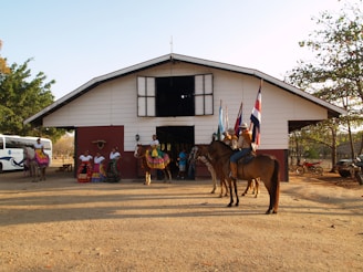 A group of people and horses are gathered in front of a ranch-like building. Several women are dressed in traditional colorful dresses, while others are wearing cowboy hats and riding horses. Flags from various countries are being held by individuals on horseback. The scene takes place in an outdoor setting with trees and a vehicle visible in the background.