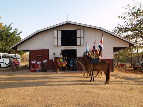 A group of people and horses are gathered in front of a ranch-like building. Several women are dressed in traditional colorful dresses, while others are wearing cowboy hats and riding horses. Flags from various countries are being held by individuals on horseback. The scene takes place in an outdoor setting with trees and a vehicle visible in the background.