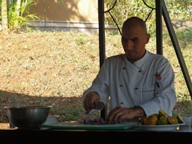 A chef in a white uniform is slicing vegetables on a cutting board outdoors. The workspace includes a metal mixing bowl and a plate with various citrus fruits. The background features a grassy slope with some greenery and a fence.