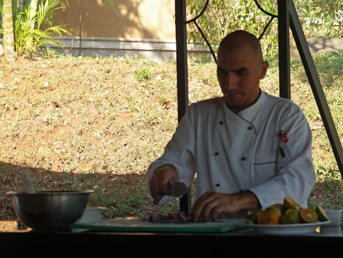A chef in a white uniform is slicing vegetables on a cutting board outdoors. The workspace includes a metal mixing bowl and a plate with various citrus fruits. The background features a grassy slope with some greenery and a fence.