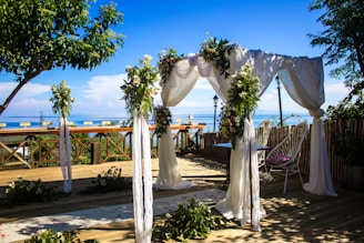 A beautifully decorated outdoor wedding arch is set up on a wooden deck overlooking a serene body of water. The arch is adorned with white fabric draping and lush greenery, complemented by floral arrangements. In the background, the blue sky and calm water create a picturesque backdrop. Chairs and tables are arranged under and around the arch, with some greenery scattered on the floor.