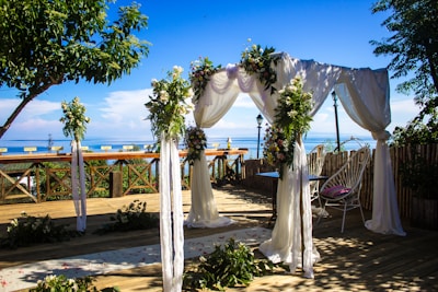 A beautifully decorated outdoor wedding arch is set up on a wooden deck overlooking a serene body of water. The arch is adorned with white fabric draping and lush greenery, complemented by floral arrangements. In the background, the blue sky and calm water create a picturesque backdrop. Chairs and tables are arranged under and around the arch, with some greenery scattered on the floor.