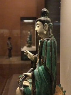 A close-up view of a statue depicting a seated figure with a serene expression. The figure wears a green robe and ornate earrings, and has a topknot hairstyle. In the background, another statue is similarly displayed, with a person observing the artifacts.