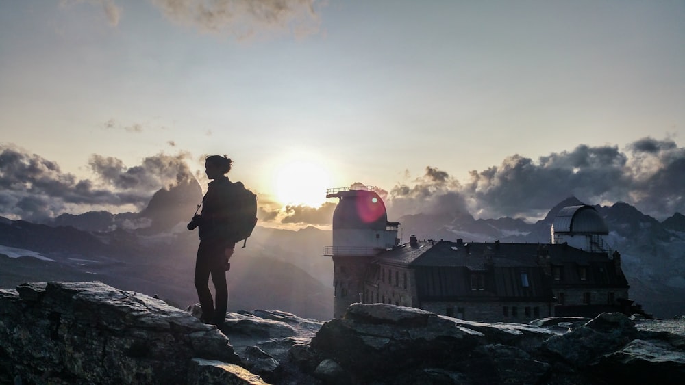 Silhouette einer Frau mit Rucksack auf einem Felsen in der Nähe eines braunen Gebäudes bei Sonnenuntergang