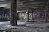 A deserted, graffiti-covered room with concrete pillars and a ceiling, featuring various colorful graffiti art on the walls. The floor is littered with debris, and the setting has an abandoned, urban decay feel.