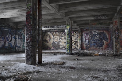 A deserted, graffiti-covered room with concrete pillars and a ceiling, featuring various colorful graffiti art on the walls. The floor is littered with debris, and the setting has an abandoned, urban decay feel.