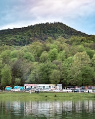 A scenic landscape featuring a lush, green forested mountain in the background with a row of trees at the base. In the foreground, there is a calm body of water reflecting the greenery. Along the shoreline, small shops and stalls with colorful flags are visible, along with several parked cars and people gathered in small groups.