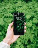 A hand holds a smartphone with a leafy green wallpaper. The background is filled with lush green ferns, creating a natural and harmonious setting.