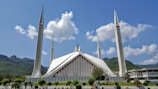 A large, modern mosque with unique architecture featuring sharp, angular lines and four tall minarets. The structure is set against a backdrop of green hills and a bright blue sky with scattered clouds. The front yard is well-maintained with greenery and a few people walking.