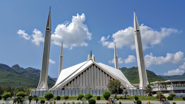 A large, modern mosque with unique architecture featuring sharp, angular lines and four tall minarets. The structure is set against a backdrop of green hills and a bright blue sky with scattered clouds. The front yard is well-maintained with greenery and a few people walking.