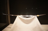 A sleek, traditional sword displayed elegantly on a white stand, with a dark background highlighting its shiny blade.