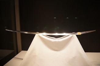 A sleek, traditional sword displayed elegantly on a white stand, with a dark background highlighting its shiny blade.