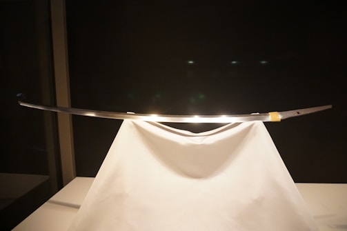 A sleek, traditional sword displayed elegantly on a white stand, with a dark background highlighting its shiny blade.