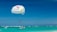 A parasailing activity is taking place over a turquoise ocean with two people suspended in a parachute attached to a speedboat. The sky is clear and blue, and the boat is skimming across the water filled with a small group of people.
