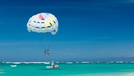 A parasailing activity is taking place over a turquoise ocean with two people suspended in a parachute attached to a speedboat. The sky is clear and blue, and the boat is skimming across the water filled with a small group of people.