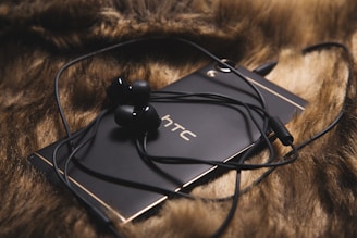 A black smartphone with the HTC logo is placed on a brown furry surface. Black wired earphones are resting on the phone, with the cable looping around it.
