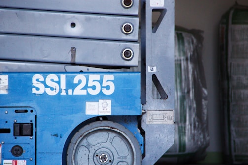 A close-up view of industrial machinery with a prominent blue color, featuring the number SSI.255 in large white text. Visible are several metallic components, including a wheel and bolts, with some wear and rust.