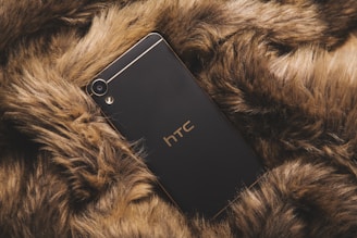 A sleek, black HTC smartphone is elegantly placed on a cozy, brown fur surface. The phone features gold accents and the brand logo is prominently visible.