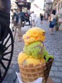 A vibrant ice cream cone with three scoops featuring bright colors such as orange, green, and purple is held up close to the camera. In the background, a cobblestone street lined with old-fashioned buildings and a horse-drawn carriage can be seen, along with a few people walking.