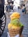 A vibrant ice cream cone with three scoops featuring bright colors such as orange, green, and purple is held up close to the camera. In the background, a cobblestone street lined with old-fashioned buildings and a horse-drawn carriage can be seen, along with a few people walking.