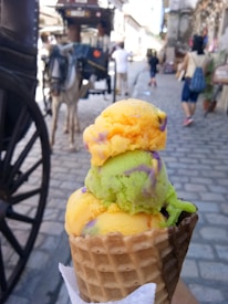 A vibrant ice cream cone with three scoops featuring bright colors such as orange, green, and purple is held up close to the camera. In the background, a cobblestone street lined with old-fashioned buildings and a horse-drawn carriage can be seen, along with a few people walking.