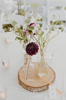 A rustic decorative arrangement with glass bottles holding pink and white flowers sits on a wooden slice centerpiece. Surrounding the centerpiece are small glass votive candle holders with lit candles.