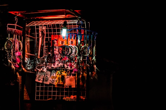 A display of various items arranged on a metal grid at a market stall, illuminated by a bright red and white light. The items include dreamcatchers with colorful feathers, various hanging trinkets, packaged goods, and other small accessories.