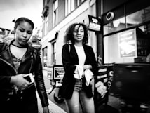 Two women are walking on a street in an urban setting. One woman is holding a smartphone and wears a leather jacket, while the other has curly hair and wears a casual outfit with a blazer. Buildings and signs are visible in the background.