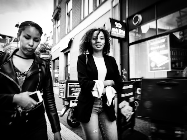 Two women are walking on a street in an urban setting. One woman is holding a smartphone and wears a leather jacket, while the other has curly hair and wears a casual outfit with a blazer. Buildings and signs are visible in the background.