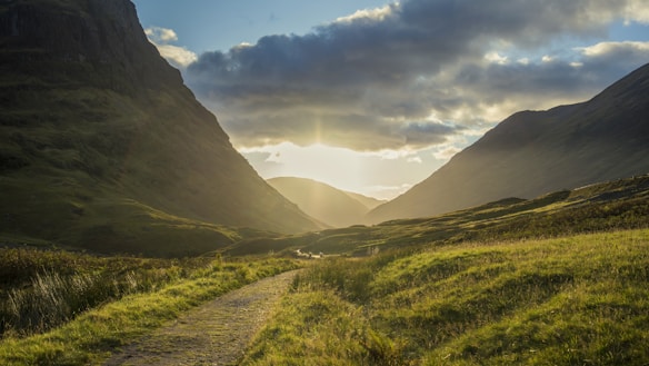 A serene mountain valley at the golden hour with a dirt path winding through lush green fields. The sun is setting behind the hills, casting a warm glow across the landscape. Thick clouds partly obscure the sky, adding depth and contrast to the scene.