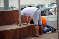 Two people are leaning forward on a wooden platform. One person is dressed in a white shirt, gray shorts, and sandals, while the other person is in an orange shirt and jeans. Several parked cars can be seen in the background.