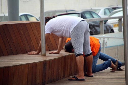 Two people are leaning forward on a wooden platform. One person is dressed in a white shirt, gray shorts, and sandals, while the other person is in an orange shirt and jeans. Several parked cars can be seen in the background.