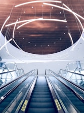 A modern escalator ascends towards a ceiling adorned with curved, illuminated light fixtures. The ceiling features a circular wooden panel that contrasts with the sleek glass and metal elements of the escalator.